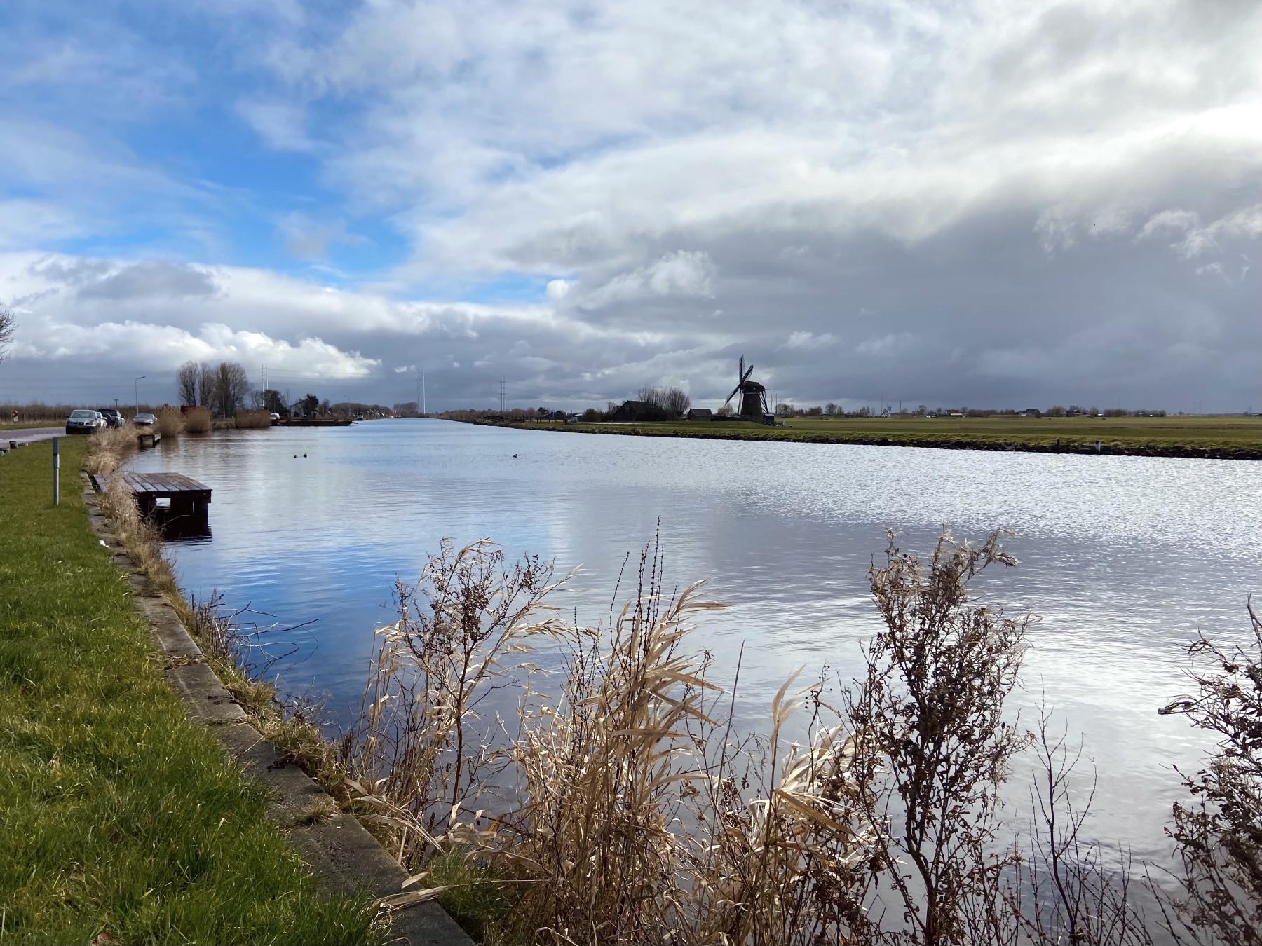 a photo of a canal under a partially sunny partially stormy sky with farmland and a windmill